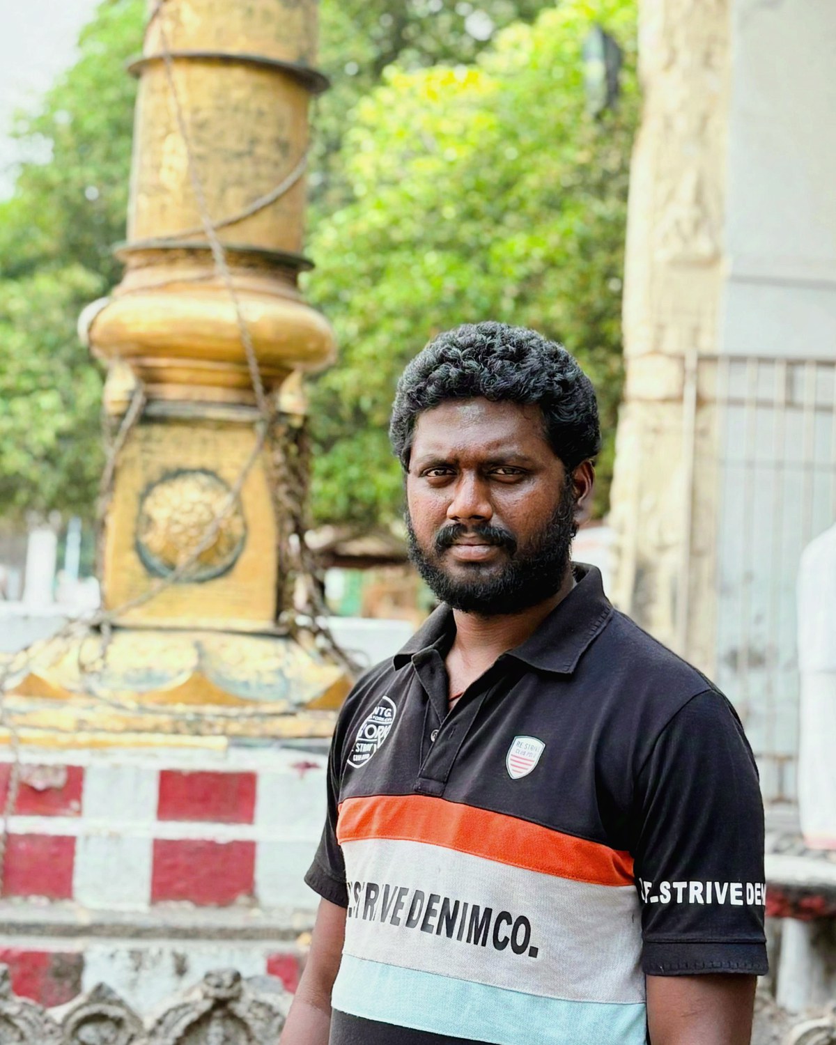 Dhanunjaya M, bearded, in a dark polo shirt, standing in a temple courtyard. Behind him a gold-clad dhvajastambha rises from a red and white plinth, with carved stone pillars and a mandapam roof to the right.
